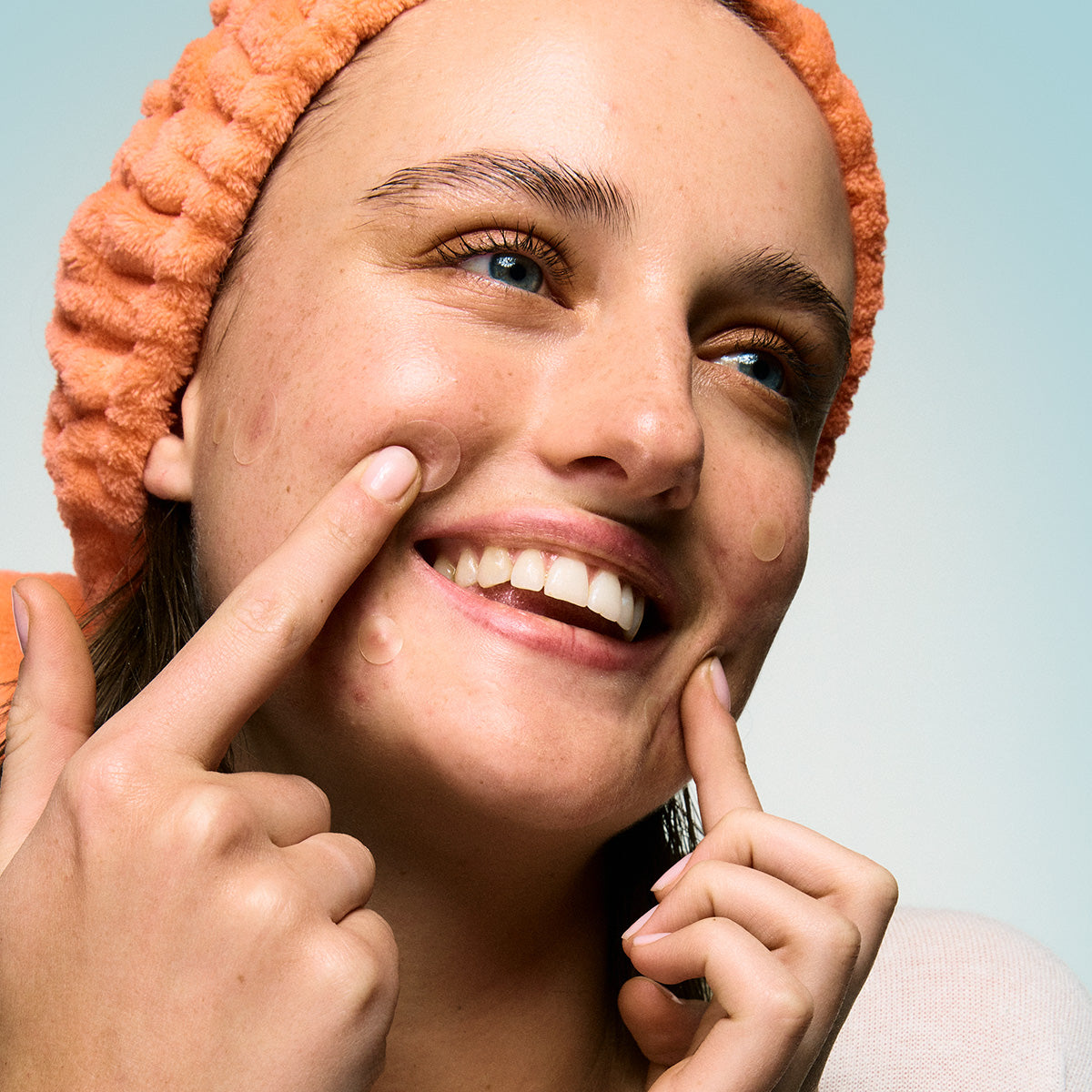 Smiling person with a peach headband applies 24/7 Zit Attack Bundle patches to their cheek with both hands.