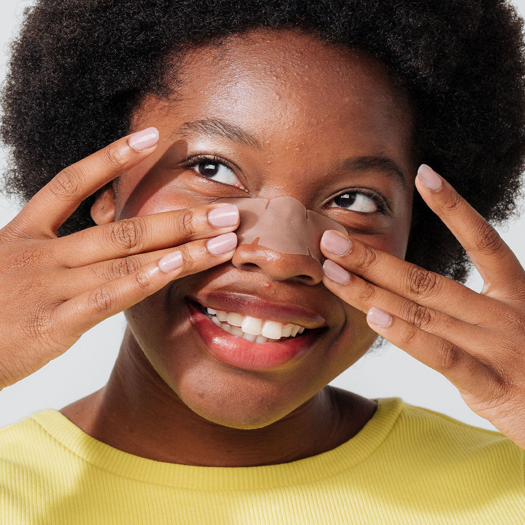 A smiling woman in a yellow top applies the GOO GETTER™ Nose strip to target blackheads.