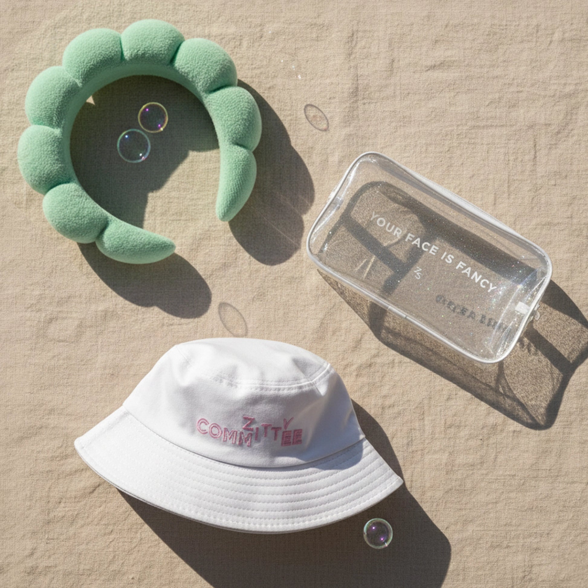 #ZittyCommittee Kit: green headband, glitter make-up bag & white bucket hat in sunlight with bubbles.